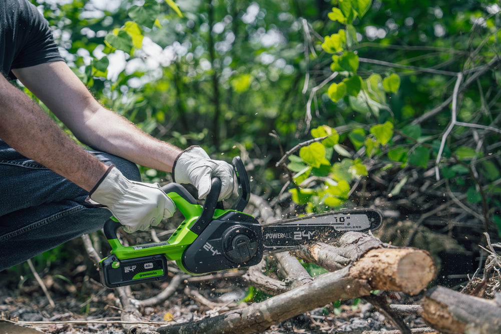 A person wearing gloves uses a green and black cordless chainsaw to cut a log outdoors in a wooded setting with green foliage in the background.