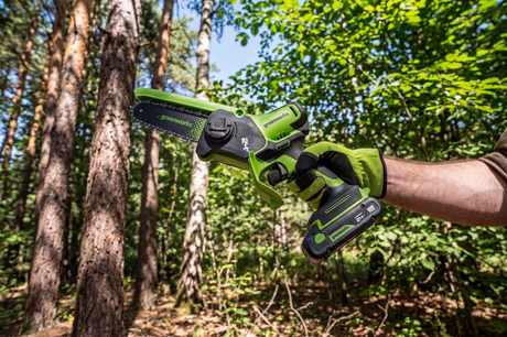 A cordless chainsaw with a green and black body is held by a person wearing a lime green and black work glove, set against a blurred forest background of pine trees.